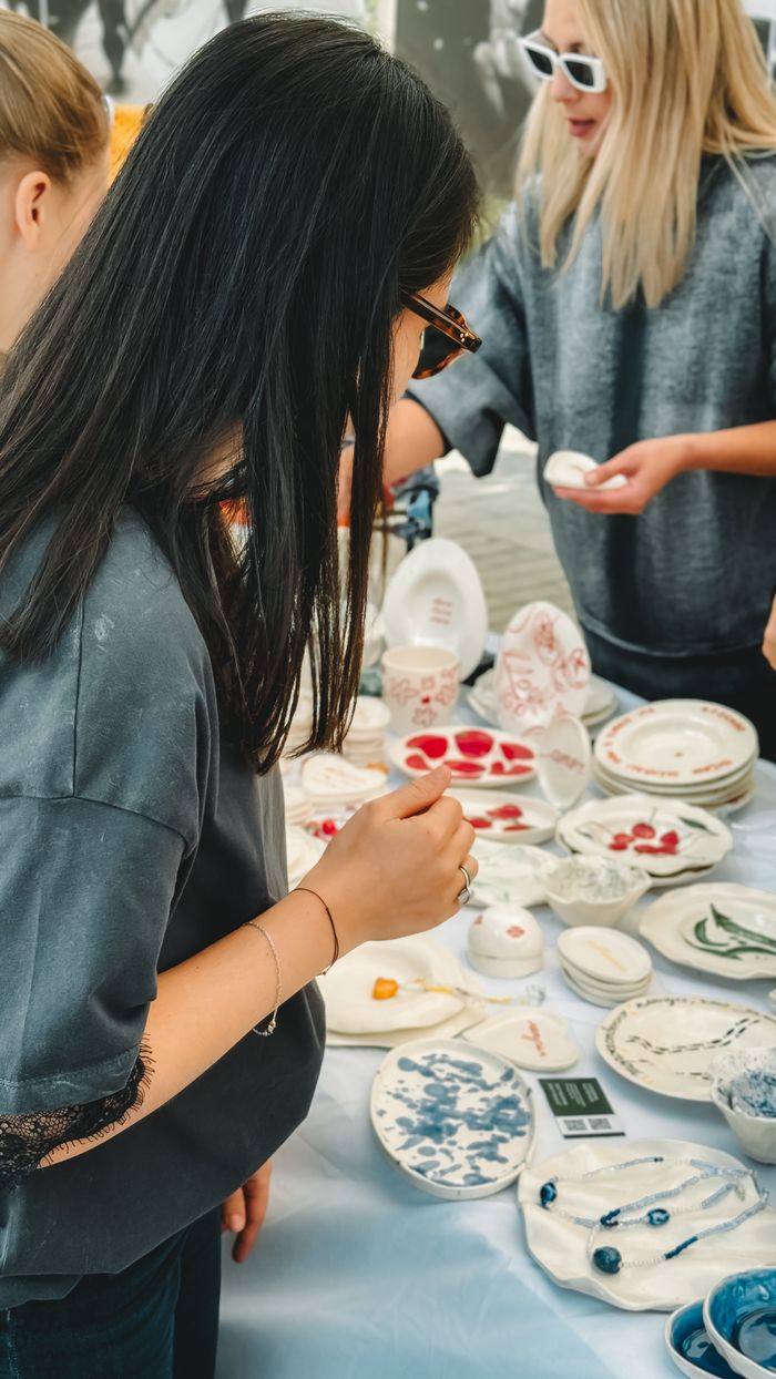 about-03 Women exploring a market stall with handmade ceramics and jewelry displayed.